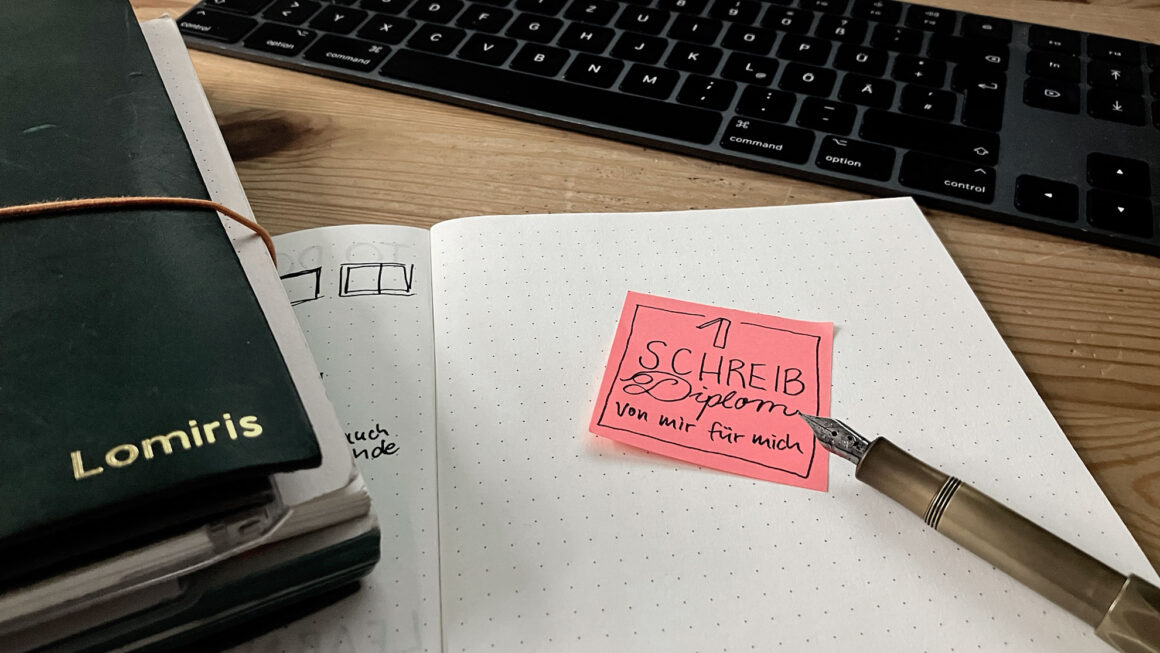 Schreibtisch mit aufgeschlagenem Notizbuch, auf dem ein selbstgemaltes „Diplom“ klebt