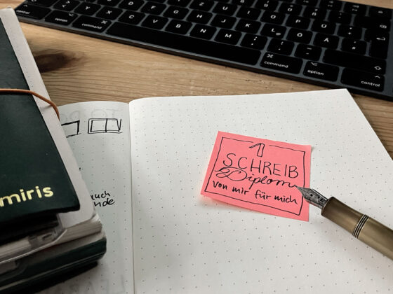 Schreibtisch mit aufgeschlagenem Notizbuch, auf dem ein selbstgemaltes „Diplom“ klebt